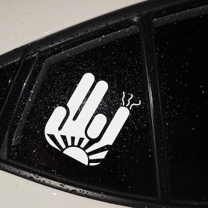 May include: White vinyl decal of a hand gesture with the index and pinky fingers extended, and the thumb and two middle fingers touching. The design includes a sunburst at the base and smoke-like lines from the pinky finger.