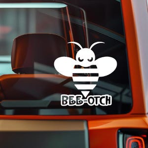 May include: White vinyl decal of a stylized bee with the text "BEE-OTCH" in a bold, sans-serif font. The bee has large wings, a striped abdomen, and a smiling face. The decal is applied to a vehicle window.