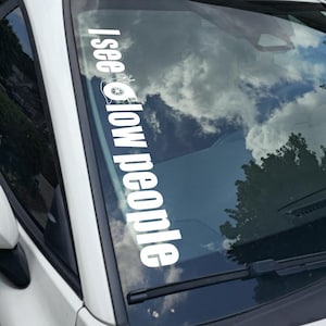May include: White vinyl car decal with the text "I see slow people" in a vertical orientation. The decal includes a small graphic of a turbocharger. The car's windshield reflects a cloudy sky and trees.