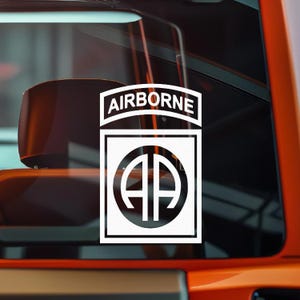 May include: White decal featuring the word "AIRBORNE" above a square emblem with a circle containing the letters "AA". The decal is displayed on a vehicle window, with the interior visible in the background.