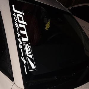 May include: White JDM sticker with Japanese text and a rising sun design, affixed to a car window. The sticker reads "JDM" vertically, with additional Japanese characters. The rising sun emblem is in a rectangular frame.