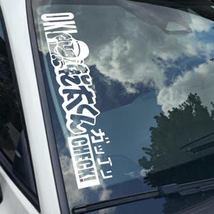 May include: A white vinyl sticker applied to a car windshield. The sticker features Japanese characters and the English word "CHECK!" in a bold, stylized font. The background shows a cloudy sky and tree reflections.