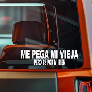 May include: White vinyl decal with the text "ME PEGA MI VIEJA PERO ES POR MI BIEN" on a car window. The decal is displayed on an orange vehicle, with a blurred background.