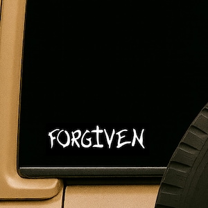 May include: White "FORGIVEN" decal on a black background, likely for a car window. The word is in a handwritten style. The image is framed by a tan vehicle panel and a portion of a black tire.