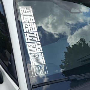 May include: White vinyl decal applied to a car windshield. The decal features vertical Japanese characters and the letters "JDM" in a bold, outlined font. The background shows a cloudy sky and a tree.