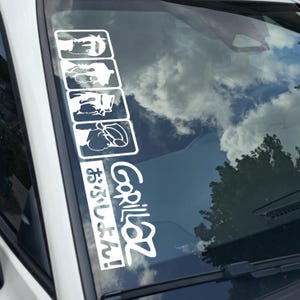 May include: White vinyl decal featuring the band Gorillaz. The decal includes the band's logo and Japanese text, along with four square illustrations of the band members. The decal is applied to a car window.