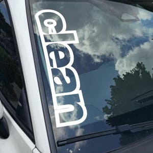 May include: White vinyl decal with the word "clean" in a bold, sans-serif font, applied to a car windshield. The word is vertically oriented. The background shows a cloudy sky and a tree.