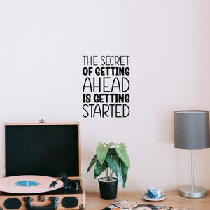 May include: A motivational quote in black text on a white wall: "THE SECRET OF GETTING AHEAD IS GETTING STARTED." A vintage record player with a pink record sits on a wooden desk, next to a potted plant and a lamp.