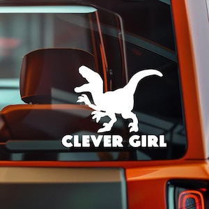 May include: White vinyl decal featuring a silhouette of a raptor dinosaur with the text "CLEVER GIRL" in a bold, sans-serif font. The decal is applied to the rear window of an orange vehicle.