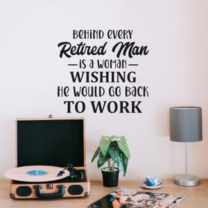 May include: A vintage wooden record player with a pink vinyl record is on a wooden table. The text on the wall says, "Behind every retired man is a woman wishing he would go back to work."