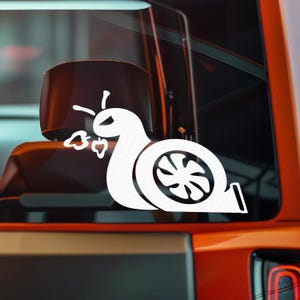 May include: White vinyl decal of a snail with a turbocharger design. The snail has a stylized eye and is expelling exhaust. The decal is applied to a vehicle window, with the vehicle's orange body visible in the background.
