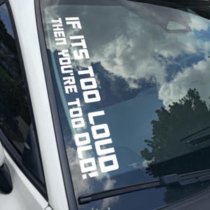 May include: White vinyl decal applied to a car windshield. The text reads: "IF IT'S TOO LOUD THEN YOU'RE TOO OLD!" The sky and trees are reflected in the glass.