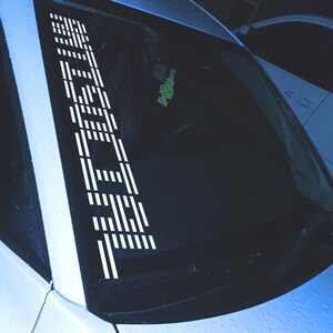 May include: White vinyl decal applied to a car windshield. The decal features the word "TECH" in a vertical, stylized font. The design is composed of a series of parallel lines and rectangular shapes, creating a modern, geometric aesthetic.