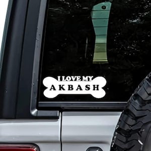 May include: A white vinyl decal applied to a vehicle window, shaped like a dog bone with the text I LOVE MY AKBASH printed in bold black capital letters. The design features a classic bone silhouette with rounded ends and a centered text layout.