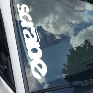 May include: White vinyl decal with the word "scrape" in a bold, sans-serif font, applied to a car windshield. The background shows a reflection of a cloudy sky and a tree.