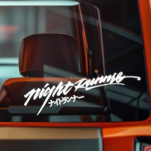 May include: White vinyl decal with the words "Night Runner" in a stylized font, with Japanese characters below. The decal is affixed to a vehicle window, with the interior visible in the background. The vehicle is orange.