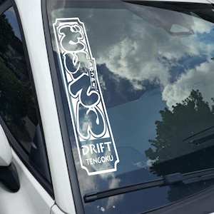May include: White vinyl sticker applied to a car windshield. The sticker features Japanese characters and the English words "DRIFT TENGOKU" in a stylized font. The background shows a cloudy sky and tree.