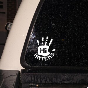 May include: White vinyl decal featuring a hand with the text "Hi Haters". The hand has five fingers and is in a waving gesture. The decal is applied to a vehicle window.
