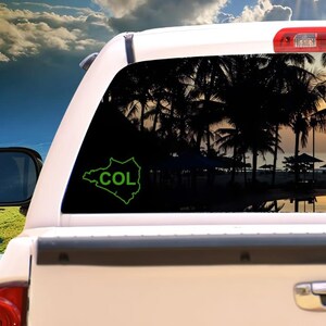 May include: A green outline of the country of Colombia with the abbreviation "COL" on the rear window of a white vehicle. The background shows a beach scene with palm trees and a sunset.
