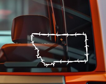 Puede incluir: Una calcomanía de vinilo blanco del estado de Montana, delineada con un diseño de alambre de púas. La calcomanía se muestra en la ventana de un vehículo naranja, con el interior visible en el fondo.