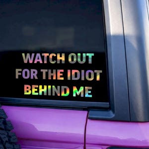 May include: A holographic sticker on a car window with the text "WATCH OUT FOR THE IDIOT BEHIND ME". The sticker has a rainbow color effect and is placed on a black background.