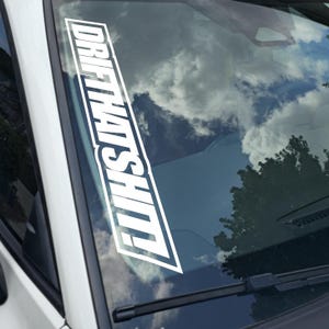 May include: White rectangular car window decal with the text "DRIFT THAT SHIT" in bold, stylized font. The decal is positioned on a car windshield, with a sky and tree visible through the glass.