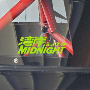 May include: A close-up of a car's interior, featuring a neon green sticker with Japanese characters and the English word "MIDNIGHT." The sticker is on a window, with red metal bars visible in the background.