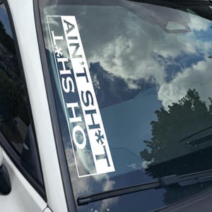 May include: White rectangular car decal with the text "AIN'T SH*T L*HS HO" in a bold, sans-serif font. The decal is affixed to a car windshield, with a sky and tree visible through the glass.