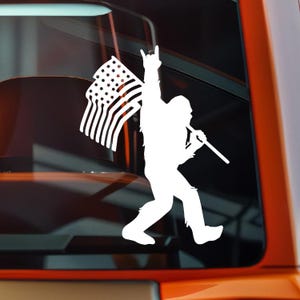 May include: White vinyl decal of Bigfoot holding an American flag and a stick, with one arm raised in a rock-and-roll gesture. The flag has stars and stripes. The design is a silhouette.
