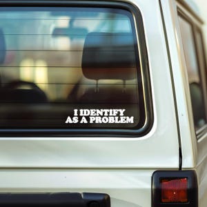 May include: White vinyl car decal with the text "I IDENTIFY AS A PROBLEM" in a bold, sans-serif font. The decal is affixed to the rear window of a white vehicle, with the interior visible through the glass.