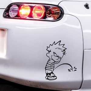 May include: A black vinyl decal of a cartoon boy urinating, affixed to the white body of a car. The boy is wearing a striped shirt and sneakers. The car's taillights are visible above the decal.