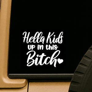 May include: White vinyl decal with the text "Hella Kids up in this Bitch" in a playful font, with a heart symbol at the end. The decal is displayed on a black background.