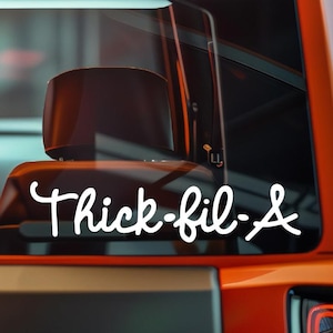 May include: White vinyl decal with the text "Thick-fil-&" in a cursive font, applied to the rear window of an orange vehicle. The decal is centered and clearly visible against the dark interior.