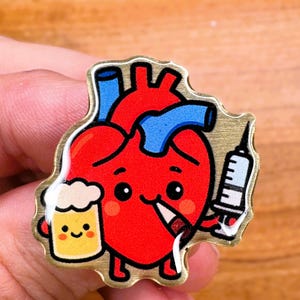 May include: A gold-colored enamel pin featuring a cartoon heart with a smiling face, holding a beer mug and a syringe. The heart is red with blue veins and a white outline.