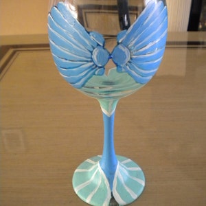 May include: A clear glass wine glass with a blue and green painted design of seashells. The design is on the bowl of the glass and the stem.