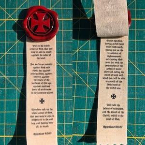 Puede incluir: Dos marcapáginas de tela con sellos de cera rojos. Un sello presenta un diseño de cruz. El texto de Efesios está impreso en los marcapáginas. Los marcapáginas están sobre una superficie de cuadrícula verde.