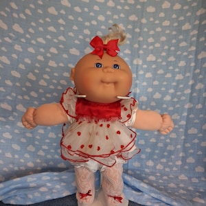 May include: A doll with a round head, blue eyes, and blonde hair tied with a red bow. The doll wears a white dress with red heart patterns, a red satin bodice, and white lace leggings with small red bows at the ankles, standing on a white base.