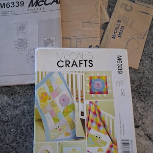 May include: McCall's Crafts M6339 sewing pattern envelope and instruction sheets for baby items. The white envelope features illustrations of a patchwork quilt, a bib, and wall art with animal motifs, alongside the text Sew Sweet Chic.
