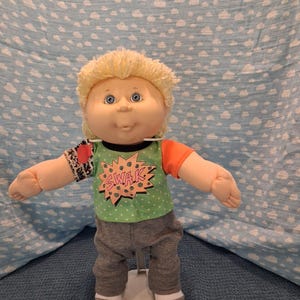 May include: A doll with blonde yarn hair and blue eyes wearing a green t-shirt with a pink starburst graphic containing the text SWAK, a black and white patterned sleeve on the right, an orange sleeve on the left, and gray pants with white socks.
