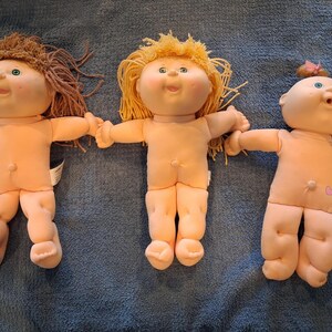 May include: Three dolls with soft fabric bodies and yarn hair. The left doll has brown yarn hair, the center doll has yellow yarn hair, and the right doll has a small brown yarn ponytail with a pink bow. All dolls have green eyes and peach skin.