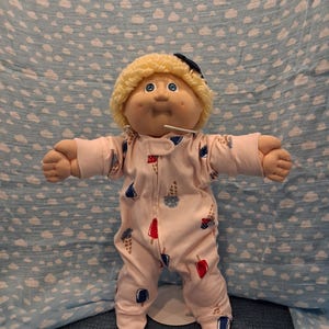 May include: A doll with blonde yarn hair and blue eyes wearing a light pink long-sleeved footed sleeper. The sleeper features a repeating pattern of colorful ice cream cones and popsicles. The doll has a round face with dimples.
