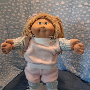 May include: A doll with tan skin, green eyes, and light brown yarn hair styled with a pink ribbon. The doll wears a light pink fleece top and shorts over a light blue and white striped long-sleeve shirt and leggings, paired with white shoes.