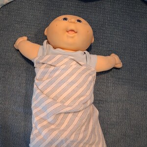 May include: A baby doll with a round, peach-toned head, blue eyes, and a small open mouth. The doll wears a light blue short-sleeved shirt and is wrapped in a white swaddle blanket featuring thin, diagonal grey stripes across its body.