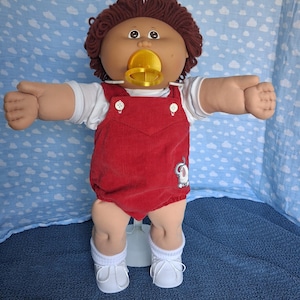 May include: A doll with brown yarn hair and a tan complexion, wearing a white short-sleeved shirt under red corduroy overalls with white buttons. The doll has a yellow pacifier in its mouth, white socks, and white shoes. An elephant patch is on the overalls.