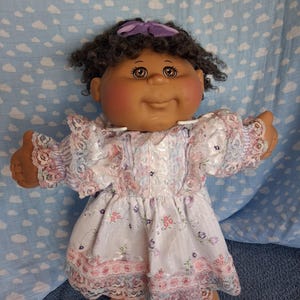 May include: A doll with dark brown curly hair, a purple hair bow, and brown eyes. The doll has a medium skin tone and rosy cheeks, wearing a white dress with a floral pattern, lace trim on the sleeves and hem, and a light purple striped bodice.
