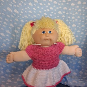 May include: A doll with light skin, blue eyes, and blonde yarn hair styled in two pigtails with red bows. The doll wears a handmade crochet dress featuring a solid pink bodice and a white skirt with a pink trim along the bottom hemline.