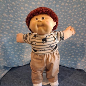 May include: A doll with a round face, dimpled cheeks, and short, curly dark red yarn hair. The doll wears a short-sleeved, grey and white striped shirt featuring black dinosaur silhouettes and tan trousers, paired with simple white shoes on its feet.