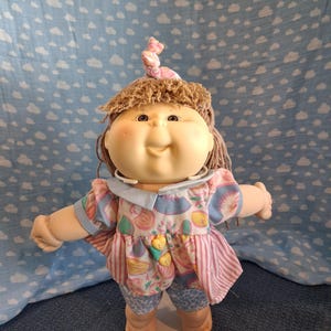 May include: A soft-bodied doll with yarn hair tied in a top knot, wearing a light blue collared shirt with a colorful fruit print, two yellow heart-shaped buttons, and blue and white patterned shorts with a pink and white striped skirt overlay.