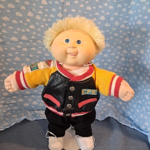 May include: A Cabbage Patch Kids doll with blonde hair and blue eyes, wearing a black and yellow varsity jacket with red trim and the letters CPK on the chest, black pants, and white shoes. The doll has a round face and a smiling expression.