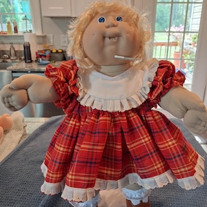 May include: A doll with blonde yarn hair and blue eyes, wearing a red plaid dress with a white ruffled collar and hem. The doll has a soft fabric body, a round face with dimples, and is dressed in white ruffled bloomers and brown shoes.